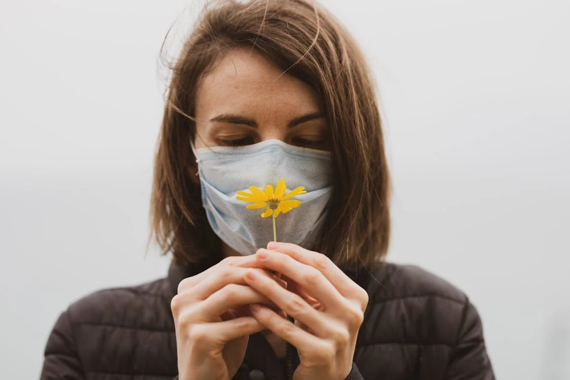 Foto di una donna con la mascherina chirurgica in faccia e una margherita gialla tra le mani