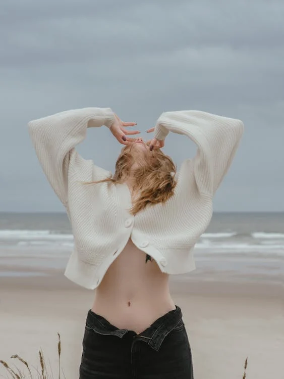 Foto di una ragazza con la pancia di fuori e un maglioncino bianco con la testa rivolta verso il cielo e il mare che si vede sullo sfondo