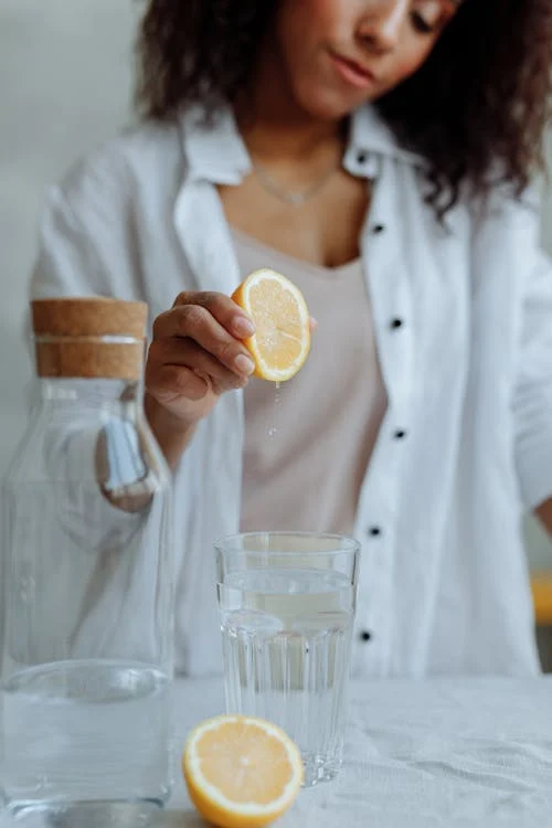 Foto di una ragazza che sta spremendo mezzo limone dentro un bicchiere d'acqua e l'altra metà del limone poggiata sul tavolo