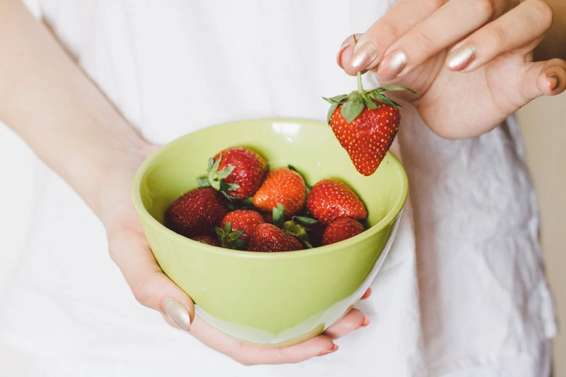 Foto di mani di donna che con una regge una ciotolina verde e l'altra prende una fragola