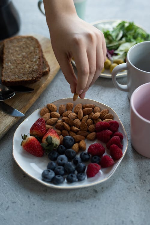 Foto di un piatto con dentro dei frutti rossi e delle mandorle e una mano che ha una mandorla in mano